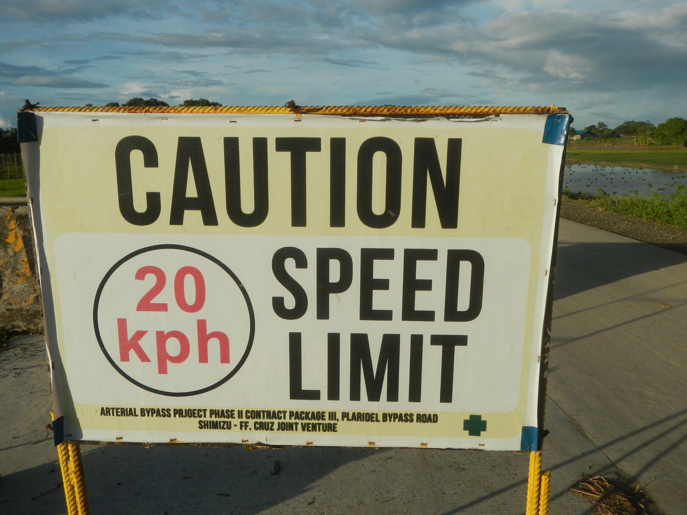 Caution speed limit sign on the side of a road with a wall, grass, water, trees, and cloudy sky in the background.