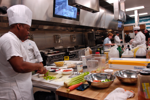 Eine Gruppe von Köchen in einer Küche beim Zubereiten von Lebensmitteln auf einem Schneidbrett, wobei ein Koch im Vordergrund in einer weißen Schürze und Mütze steht und ein Messer hält und von verschiedenen Utensilien, Schüsseln, Gemüse, einem Handy, Flaschen und anderen Gegenständen umgeben ist, mit einem Fernseher und Deckenleuchten im Hintergrund.