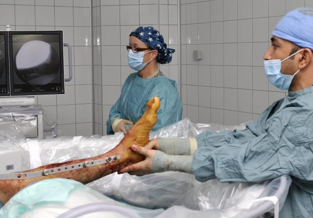 Two surgeons in scrubs and masks performing surgery on a patient's leg in an operating room with medical equipment in the background.