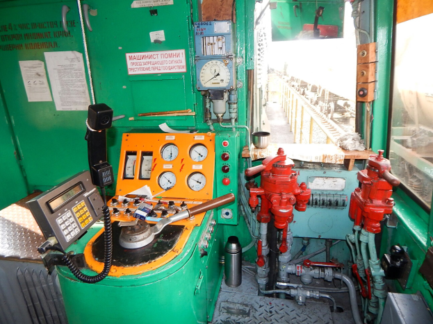 Ein Nahaufnahme-Bild eines Zugsteuerpults mit Messgeräten, Feuerhydrant, Rohren und anderen Gegenständen, mit Papieren an der linken Wand und einem Glasfenster auf der rechten Seite.