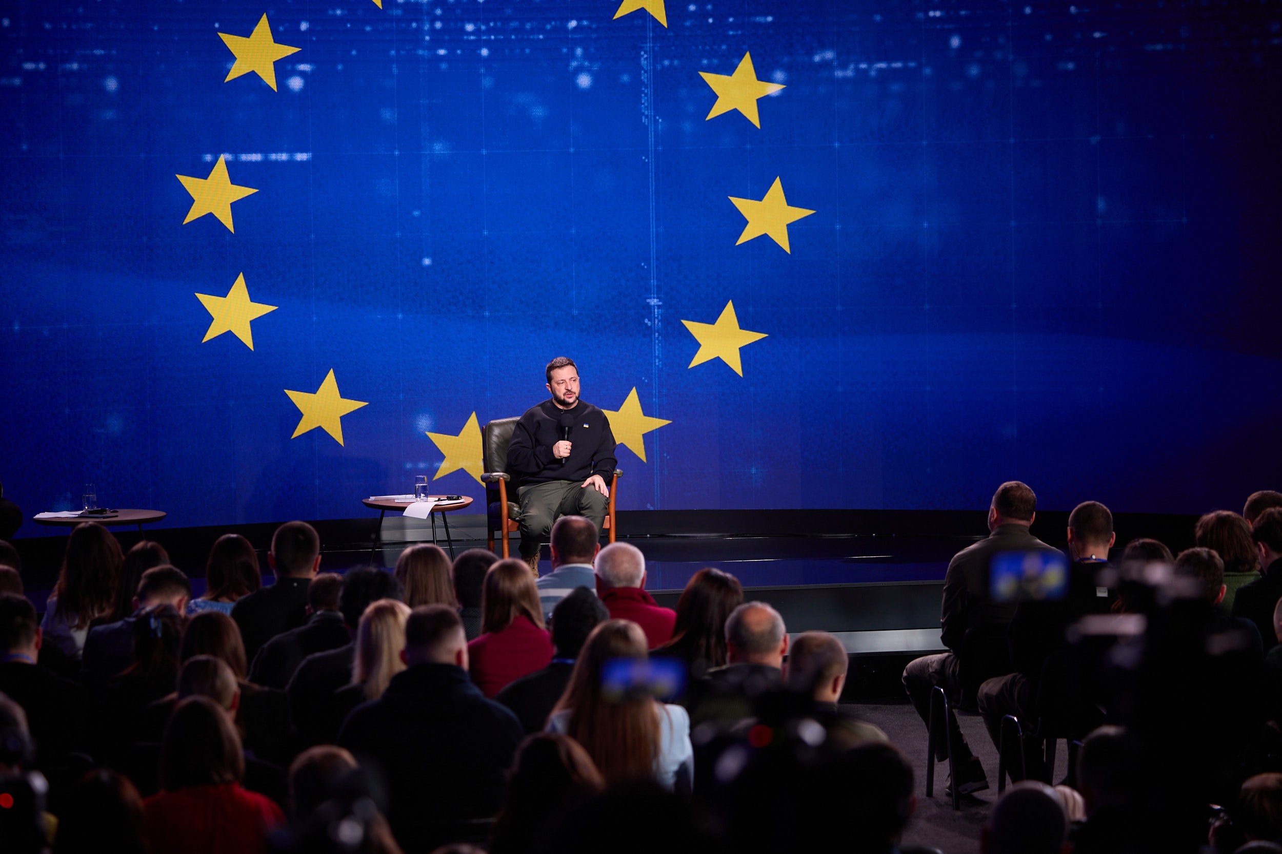 A man in a black dress sits on a stage holding a microphone, addressing a seated audience; two tables with glasses flank him, and a blue screen with yellow stars is visible behind him.