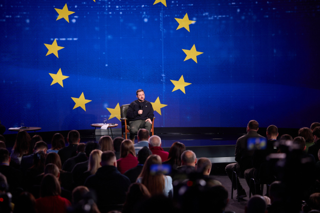 A man in a black dress sits on a stage holding a microphone, addressing a seated audience; two tables with glasses flank him, and a blue screen with yellow stars is visible behind him.