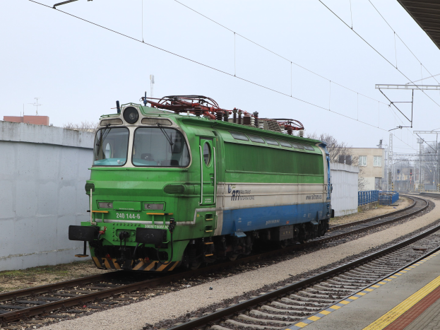 Ein grünes und blaues elektrisches Lokomotiv, das auf den Schienen neben einem Bahnsteig führt, mit einer Wand, elektrischen Masten mit Drähten, Bäumen, Gebäuden und einem klaren Himmel im Hintergrund.