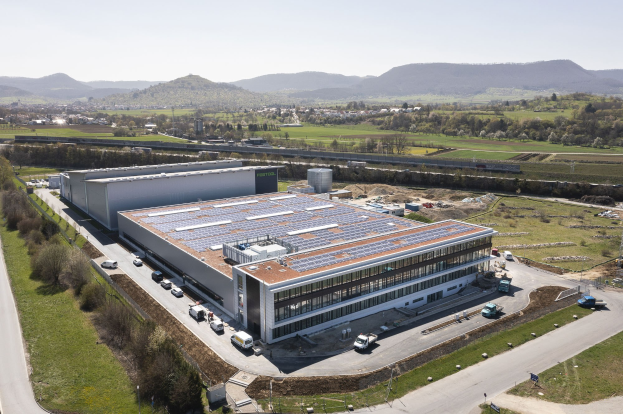Aerial view of a large building with solar panels on its roof, surrounded by vehicles, trees, grass, and poles, with hills and a clear blue sky in the background.