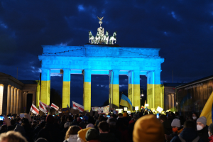 A crowd of people holding flags and placards stands in front of the Brandenburg Gate in Berlin, Germany, with a banner on the right side and cloudy skies above.