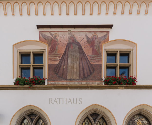 A building with windows and a mural on its side, surrounded by flowering plants, with the word "Rathaus" at the bottom.