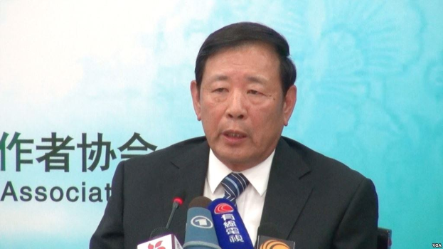 Ein Mann im Anzug sitzt an einem Tisch mit Mikrofonen und spricht während einer Pressekonferenz, mit einem Banner im Hintergrund.