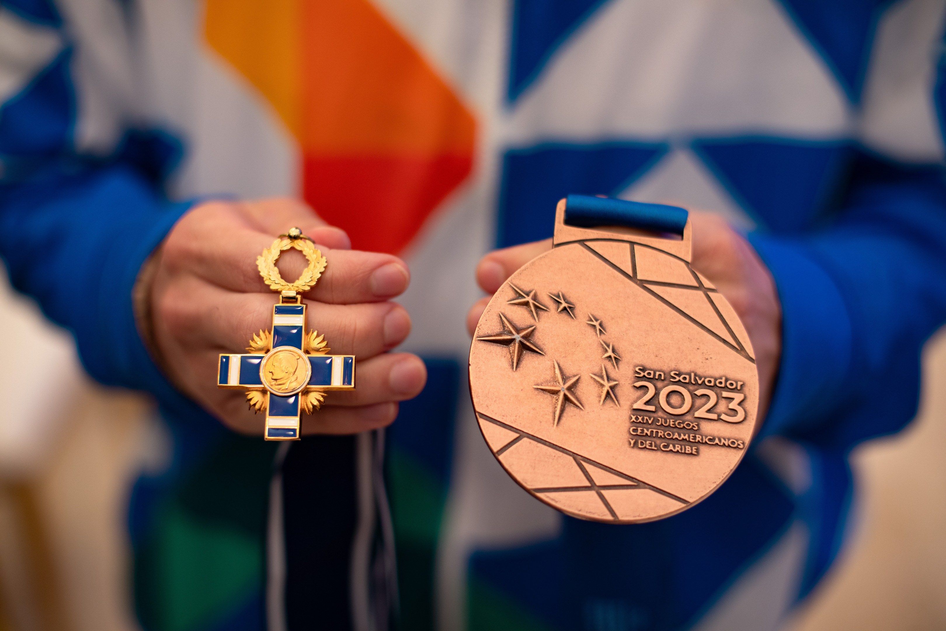 A person holding a gold medal with text reading "2023 San Salvador Centroamericano" in their hands against a blurred background.