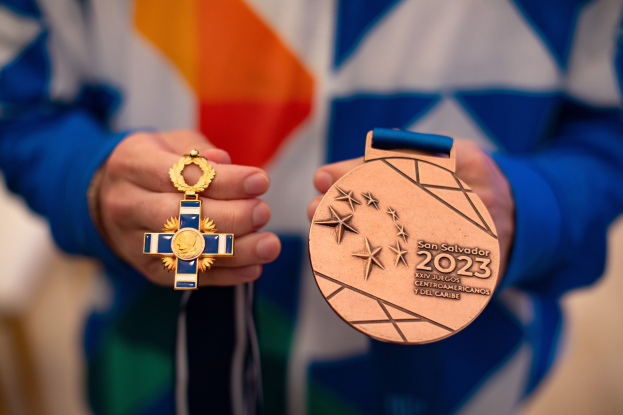 A person holding a gold medal with text reading "2023 San Salvador Centroamericano" in their hands against a blurred background.