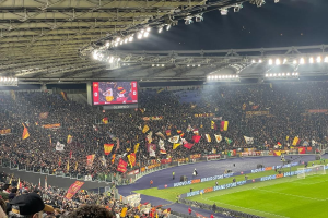Eine große Menschenmenge in einem Stadion mit schwingenden Fahnen, Gras, einem Netz, Bannern, Menschen in Mützen, einem Bildschirm und beleuchteter Decke, was auf Spannung vor einem Fußballspiel hindeutet.
