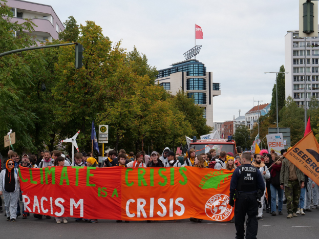 Eine Gruppe von Menschen marschiert eine von Bäumen gesäumte Straße entlang, die ein Banner mit der Aufschrift "Klima-Krise ist eine Krise" trägt, mit parkenden Fahrzeugen, Schildern und Gebäuden im Hintergrund unter einem klaren blauen Himmel.