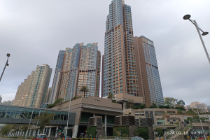 Eine Stadtstraße mit hohen Gebäuden, Fahrzeugen, Laternenmasten, einer Brücke, Bäumen und einem bewölkten Himmel, mit einem prominenten Apartmenthaus im Vordergrund.