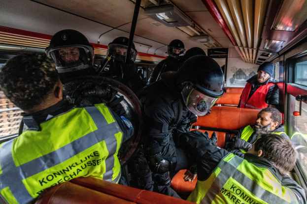 Eine Gruppe von Polizisten in Einsatzausrüstung sitzt auf einem Bus, wobei eine Person auf dem Sitz in der Mitte Platz genommen hat und ein Plakat im Hintergrund zu sehen ist.