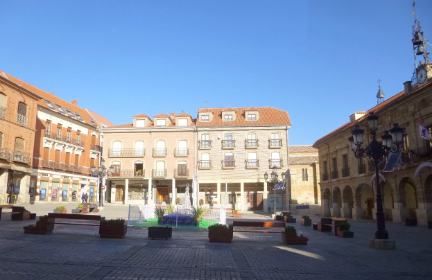 A city square with a central fountain surrounded by benches, potted plants, street poles, and lights, featuring a clock tower and buildings under a clear blue sky.