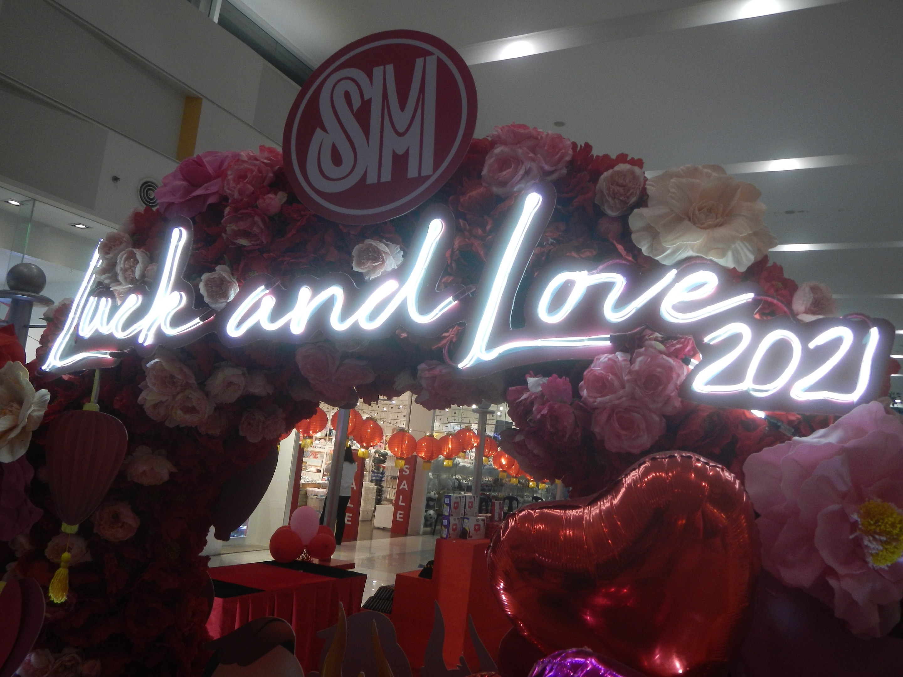 Storefront sign reading "Luck and Love 2021" decorated with flowers, balloons, and festive items during an SM Valentine's Day celebration.
