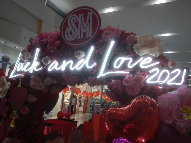 Storefront sign reading "Luck and Love 2021" decorated with flowers, balloons, and festive items during an SM Valentine's Day celebration.
