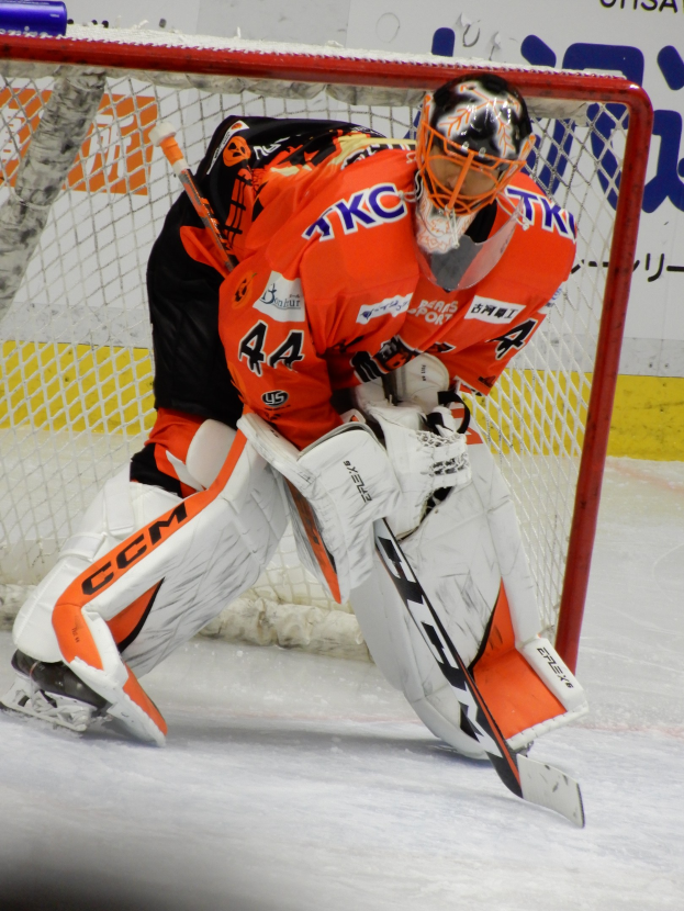 Eishockey-Spieler in orange-weißer Uniform, der vor einem Tor einen Rückschlag macht, mit Helm, Handschuhen und Hockey-Schläger, mit sichtbarem Torpfosten und einer Tafel mit Text im Hintergrund.