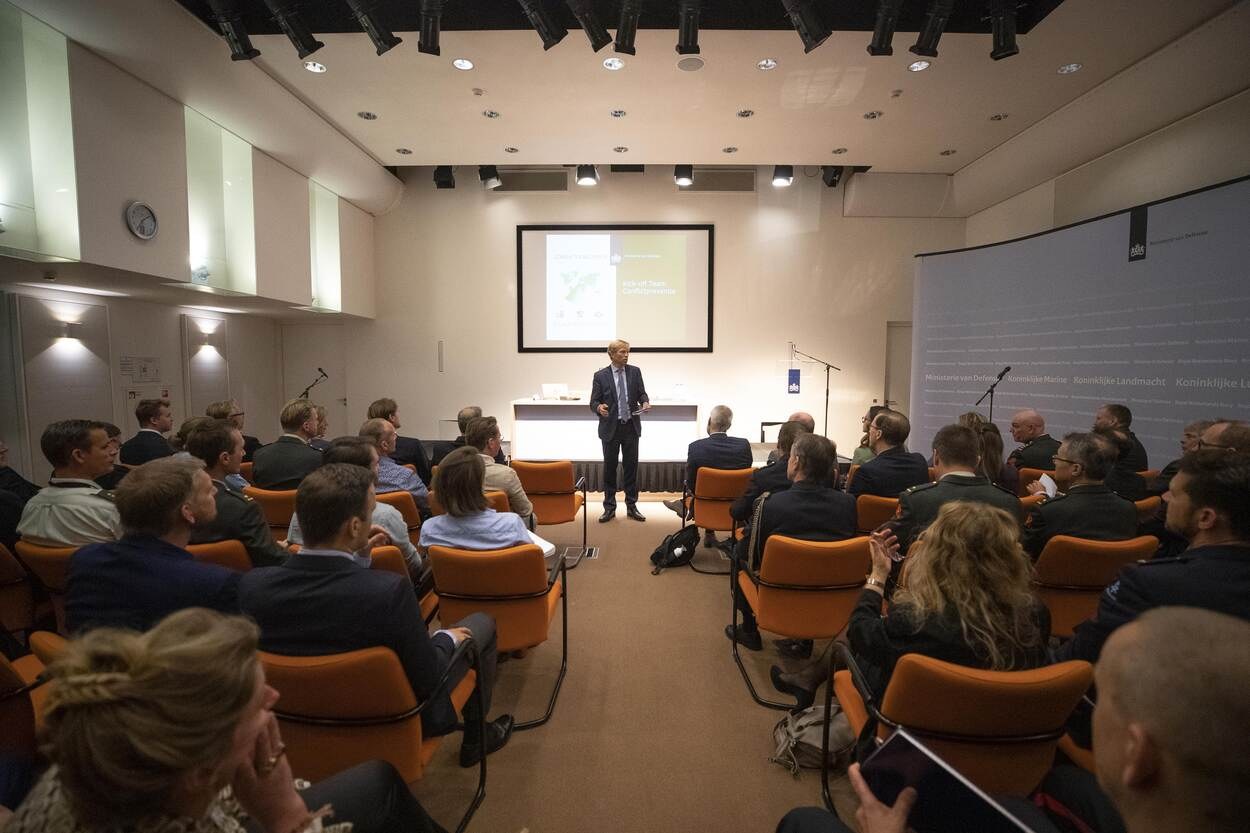 A man stands speaking into a microphone while an audience sits in chairs facing a projector screen displaying "Koninklijke Landmänch - The Future of the Future," with a clock, lights, and ceiling fixtures visible.