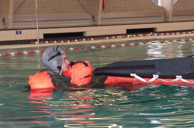 Ein Mann in einem Schwimmwesten und Helm treibt auf einem aufblasbaren Floß in einem Swimmingpool, mit einer Wand, einem Pfahl und anderen Gegenständen im Hintergrund.
