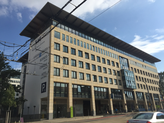 Large modern building with numerous windows, labeled "Stadthalle Center for Business," surrounded by trees, street infrastructure, vehicles, pedestrians, and a cloudy sky.