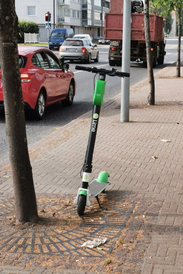 Elektroroller auf einer Stadtstraße neben einem Baum geparkt, mit Fahrzeugen und Gebäuden im Hintergrund.