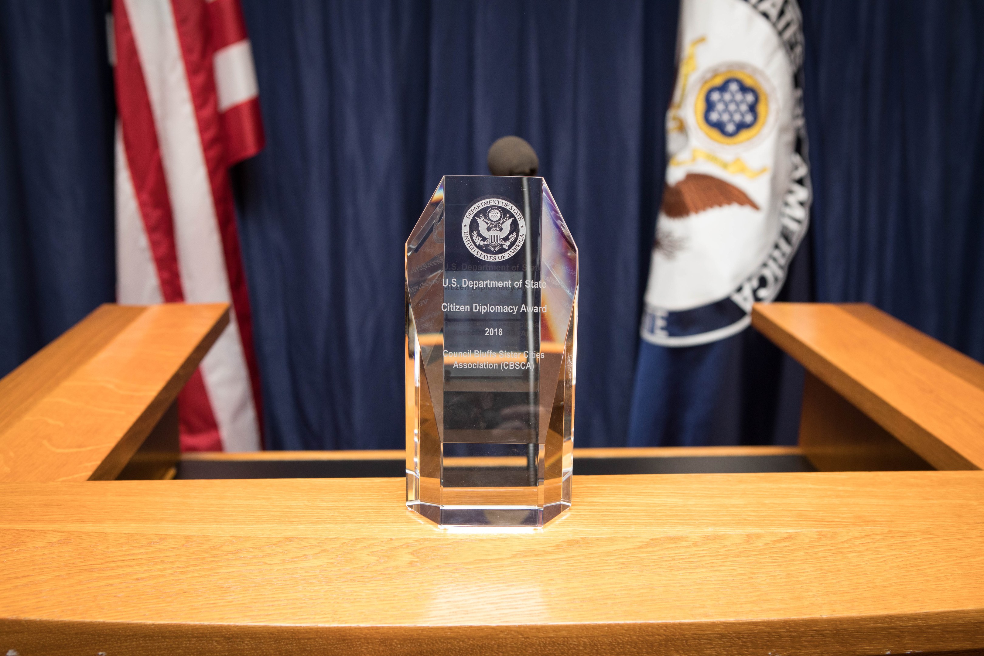 Ein Glaspreis auf einem hölzernen Podest mit einem Mikrofon davor, Flaggen und Vorhänge im Hintergrund.