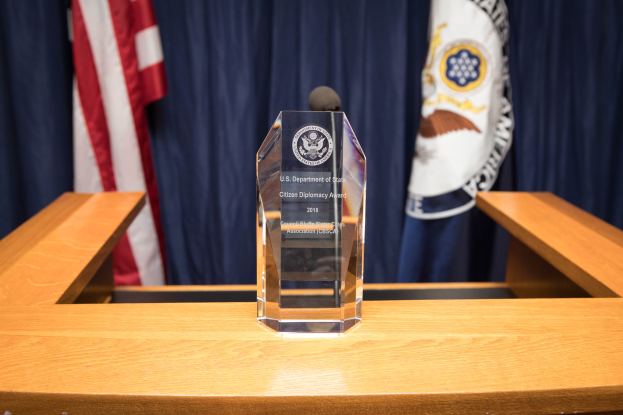 Ein Glaspreis auf einem hölzernen Podest mit einem Mikrofon davor, Flaggen und Vorhänge im Hintergrund.