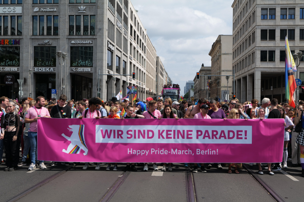 Eine Gruppe von Menschen marschiert auf einer Straße in Berlin, Deutschland, mit einer rosafarbenen 'Happy Pride March'-Fahne, mit Gebäuden, Laternenmästen und Verkehrszeichen an der Straße unter einem bewölktem Himmel.