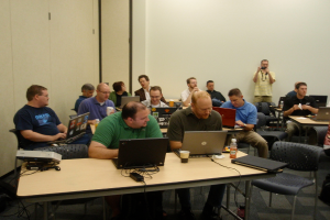 Eine Gruppe von Menschen, die auf Stühlen sitzen, wobei eine Person in einem gelben Hemd eine Kamera hält, während andere einen Laptop bedienen.