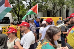 Eine Gruppe von Menschen steht vor einem Gebäude, einige tragen Mützen und Taschen und halten palästinensische Flaggen während einer Demonstration mit Fahrzeugen auf der Straße, Bäumen und einem klaren blauen Himmel im Hintergrund.
