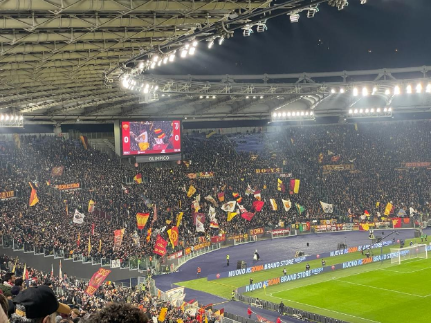 Large crowd in a stadium with waving flags, banners, and a screen, under illuminated ceiling lights, suggesting anticipation for a soccer match.
