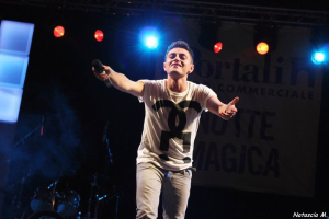 Ein Mann steht auf einer hell erleuchteten Bühne und hält ein Mikrofon in der Hand, im Hintergrund sind Musikinstrumente und ein "Nouveau Magica"-Plakat zu sehen.