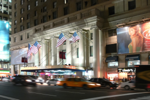 Lebendige Stadtstraße bei Nacht mit fahrenden Fahrzeugen, einem Gebäude mit Fenstern, Säulen und Fahnenmasten im Hintergrund, beleuchteten Schildern mit Text und einem Bild einer Frau sowie Straßenbeleuchtung.