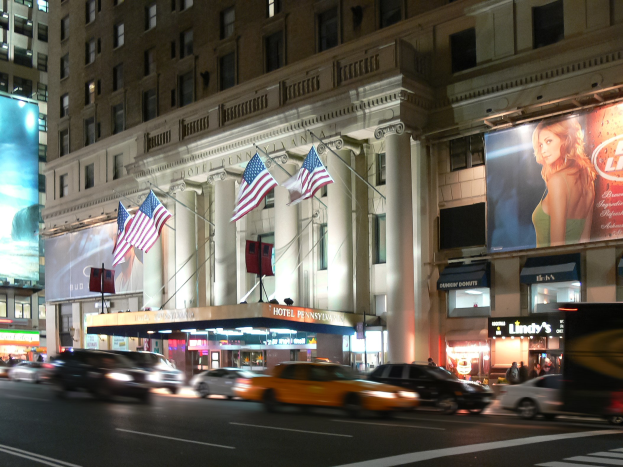 Lebendige Stadtstraße bei Nacht mit fahrenden Fahrzeugen, einem Gebäude mit Fenstern, Säulen und Fahnenmasten im Hintergrund, beleuchteten Schildern mit Text und einem Bild einer Frau sowie Straßenbeleuchtung.