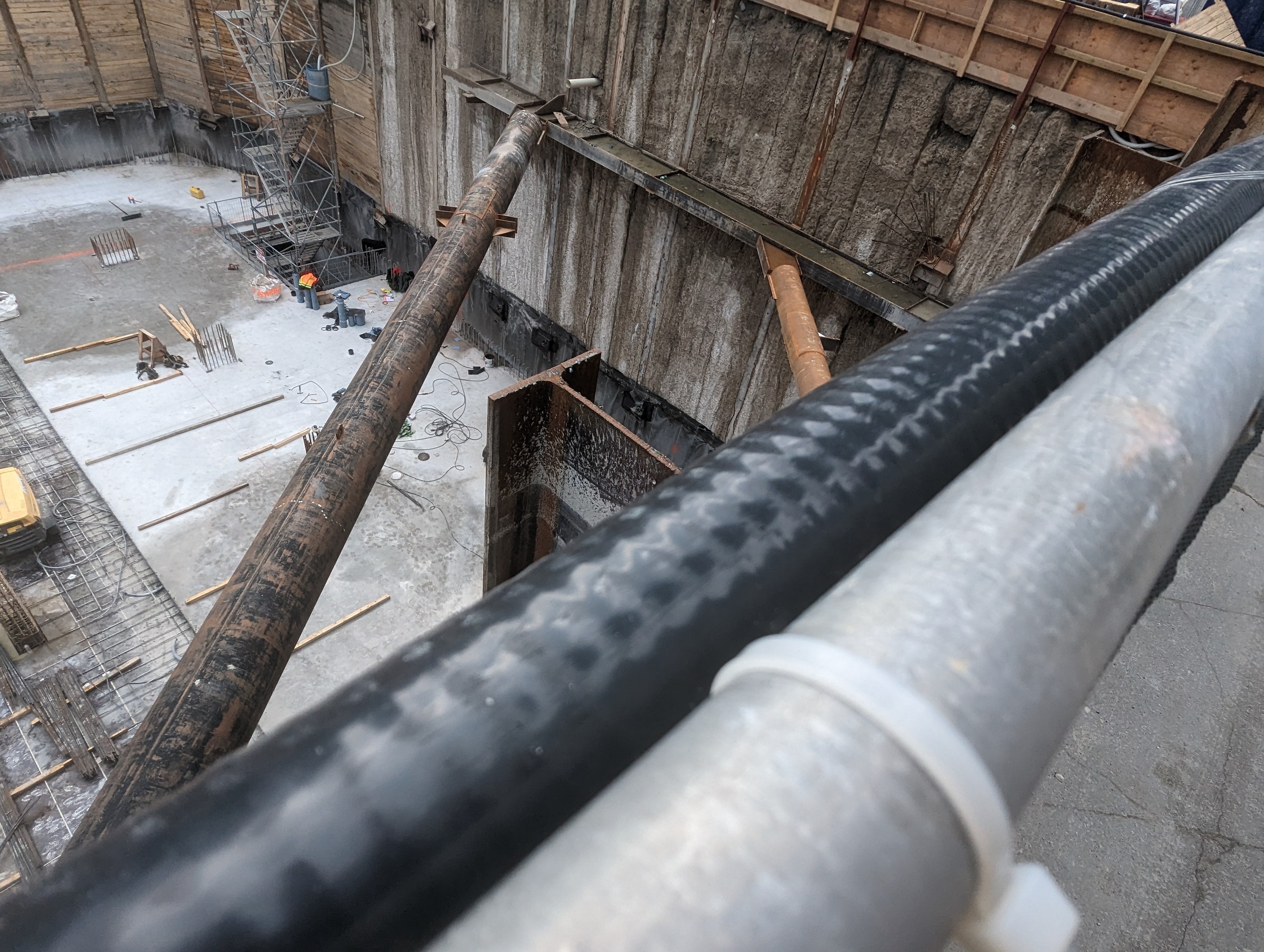 Baustelle mit einem großen Metallrohr, Menschen, hölzernen Gegenständen und Geräten auf dem Boden, Fahrzeugen und einem Gebäude im Hintergrund.