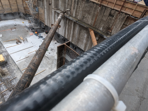 Baustelle mit einem großen Metallrohr, Menschen, hölzernen Gegenständen und Geräten auf dem Boden, Fahrzeugen und einem Gebäude im Hintergrund.