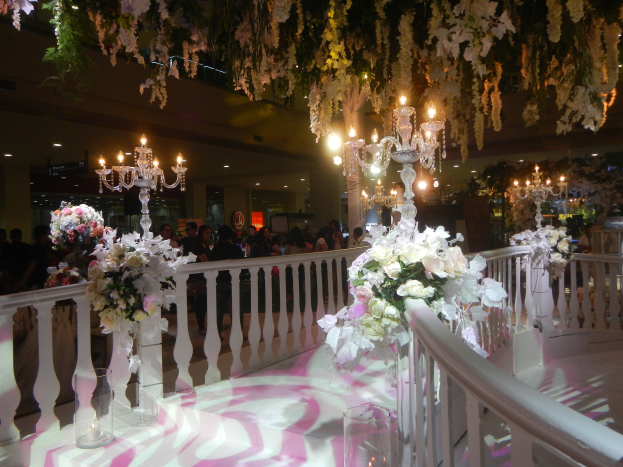A wedding ceremony in a large room decorated with white and pink flowers, chandeliers, and greenery, with guests standing in the background and floral arrangements on the floor.