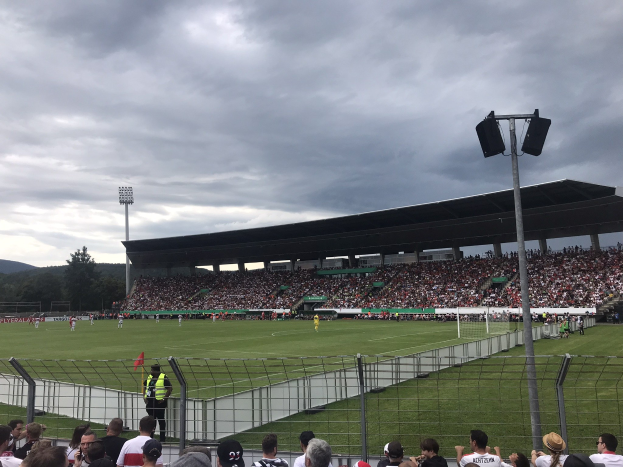 Große Menschenmenge bei einem Fußballspiel in einem Stadion mit beleuchtetem Feld, umgeben von einem Zaun, Bäumen und Hügeln im Hintergrund.