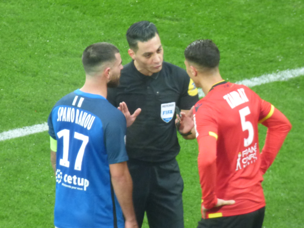 Referee in black shirt and pants speaking with two men in blue and red shirts on a soccer field, all wearing glasses.
