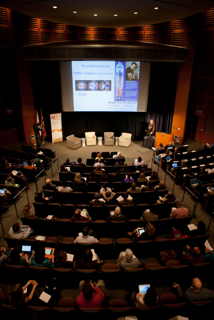 Ein mit Sitzenden Personen gefüllter Hörsaal, beleuchtet von zahlreichen Lichtern, mit Fahnen links, einem Podium rechts und einer Werbetafel links.
