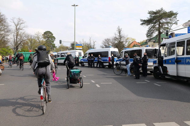 Gruppe von Menschen, die auf Fahrrädern eine Straße entlangfahren, mit Polizeiwagen, anderen Fahrzeugen, Bäumen, Laternen, Verkehrsampeln, Schildern, Gebäuden und einem bewölkten Himmel.