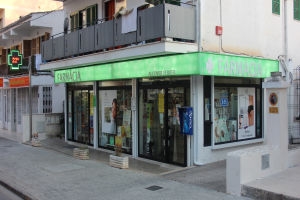 Eine Apotheke an einer Straßenecke mit Gebäuden, Geschäften, Fahrzeugen und einem klaren blauen Himmel.
