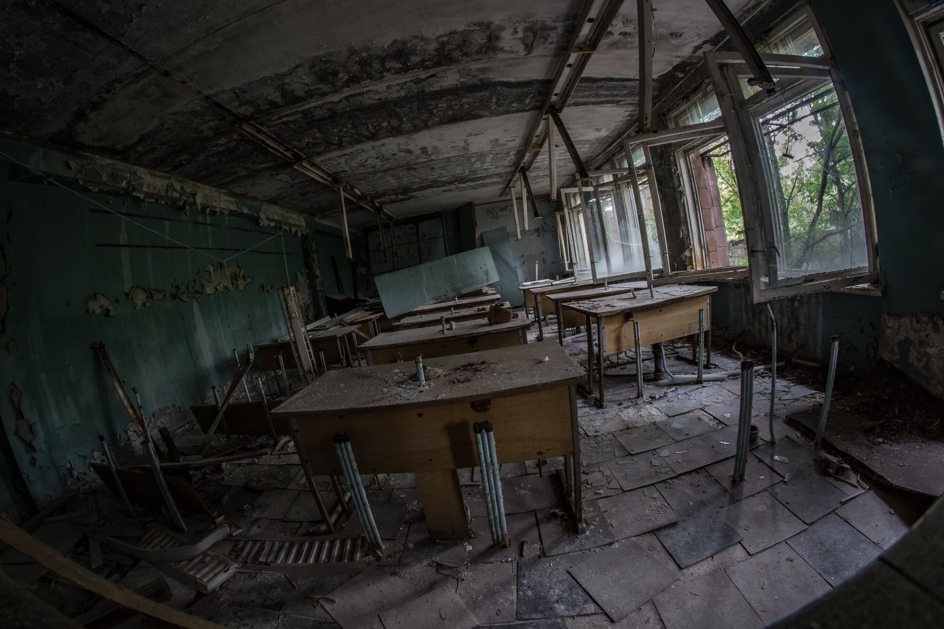Verlassener Klassenraum in Pripjat, Tschernobyl, Ukraine mit Tischen, Stühlen, abblätternden Wänden, kaputten Fenstern und sichtbarer Decke, umgeben von Bäumen draußen.