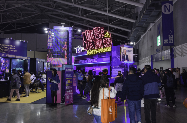 Eine große Gruppe von Menschen steht vor einem Messestand mit der Aufschrift "Indie Game Development Award 2019", einige halten Taschen und verschiedene Displays und Bildschirme sind im Hintergrund zu sehen.