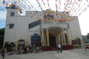 Eine Gruppe von Menschen, die Fahrräder vor einer weißen Kirche mit architektonischen Details wie Fenstern, Säulen, Bögen, Türen und einem Kreuzsymbol fahren, mit Pflanzen, einem Zaun, einer Fahne, Bäumen und einem bewölkten Himmel im Hintergrund und bunten Fahnen, die an einem Seil oben befestigt sind.
