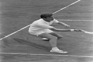 Ein Mann in wei├čer Tennisbekleidung befindet sich in einer Hockposition auf einem Tennisplatz und h├Ąlt einen Tennisschläger in der Hand, wobei er entschlossen aussieht.