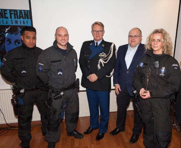 Eine Gruppe von Polizeibeamten steht vor einem Bildschirm und einem Plakat an der Wand, mit einem Tisch voller Gegenstände und Kabel auf der rechten Seite.