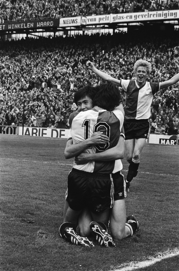Drei Fußballspieler umarmen sich auf dem Boden nach einem Tor, mit einer jubelnden Menge und schwenkenden Fahnen in einem schwarz-weißen Stadion-Setting.