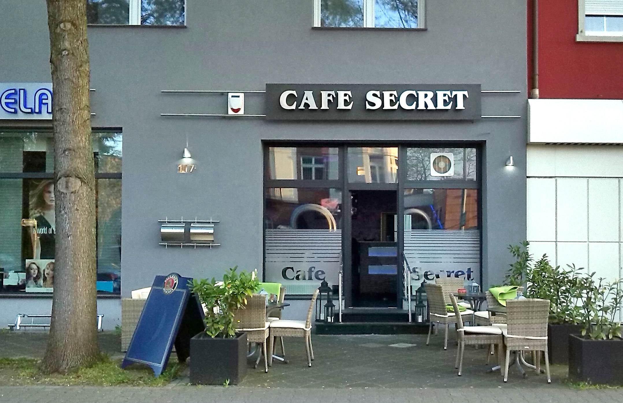 Cafe Secret in Berlin, Germany, with a building, signboard, outdoor seating, potted plants, a board, a tree, and grass.