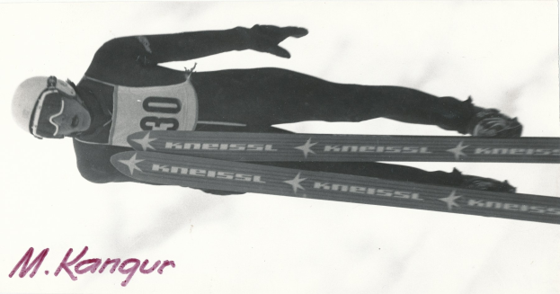 Schwarzes und weißes Foto eines Skifahrers in der Luft, der einen Helm, eine Brille, Handschuhe und Schuhe trägt und Ski hält, mit Text unten.